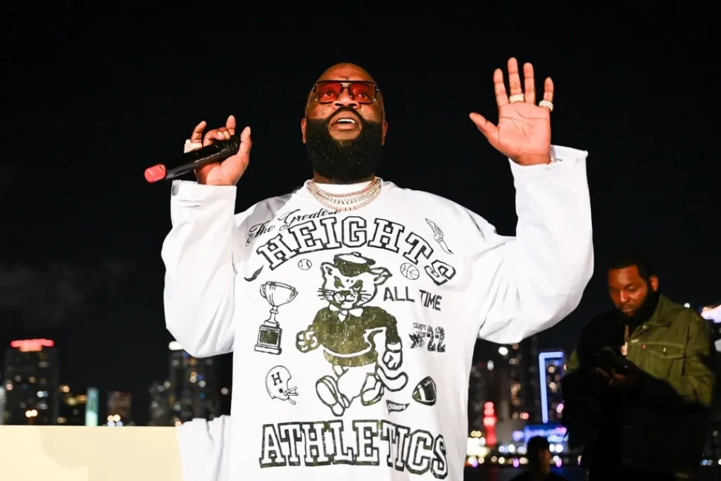 Under the night lights, Rick Ross rocks tinted aviator sunglasses and a diamond cuban link chain